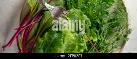 banderole avec mélange de salades dans l'assiette et à la main avec fourchette sur la table texturée blanche. Jeunes pousses juteuses de pois ou de haricots, pousses de betteraves et salade verte. Guérir Banque D'Images