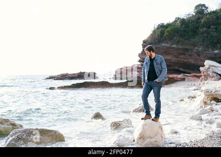 Homme en Jean et une veste en denim se tient sous la coupe ses pieds sur une grande pierre sur une plage de galets sur fond de montagnes rocheuses verdoyantes Banque D'Images
