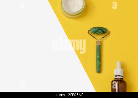 Composition créative avec rouleau de jade, crème faciale, sérum sur fond jaune et blanc de deux couleurs avec espace pour le texte Banque D'Images