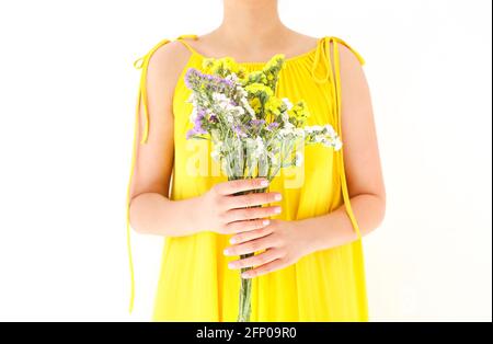 Femelle méconnaissable en robe jaune se cachant derrière un bouquet de fleurs sauvages le jour de l'été sur fond blanc Banque D'Images