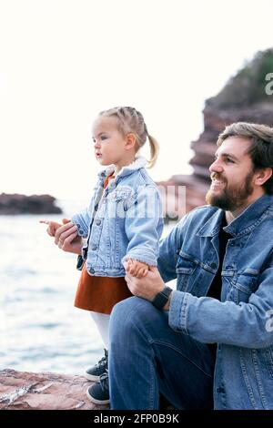 Un père souriant tient les mains d'une petite fille, pointant avec un doigt dans la distance, assis sur une côte rocheuse contre la toile de fond des montagnes et Banque D'Images