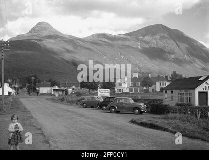 1960, historique, des voitures de l'époque garées à l'extérieur des salons de thé de Fraser dans le village de Glencoe, Highlands, Écosse, Royaume-Uni avec les montagnes écossaises de Pap de Glencoe en arrière-plan. Glen COE est l'une des plus célèbres glens des Highlands d'Écosse et est connue comme la porte d'entrée des Highlands. Banque D'Images