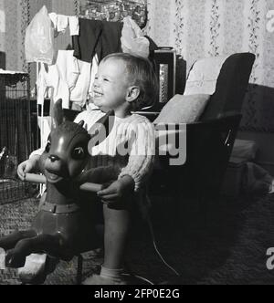 1960s, historique, à l'intérieur d'une pièce de devant, un petit garçon excité assis sur son cheval à bascule jouet. Un fireguard et quelques vêtements de séchage, avec une cage à oiseaux sur un téléviseur également dans l'image. Banque D'Images