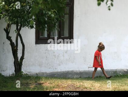 Belarus - 16,06,2007: Une fille dans une robe rouge marche près de la maison. Maison blanche, fenêtre, arbre. Banque D'Images