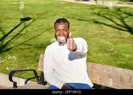 Un jeune homme afro-américain décontracté assis sur un banc de parc et montrant les pouces vers le haut geste à la caméra. Photo de haute qualité Banque D'Images