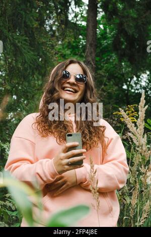 Une jeune femme dans un sweat-shirt rose, dans des lunettes de soleil, avec un téléphone, rire Banque D'Images