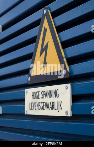 Panneau de danger électrique sur un panneau triangulaire jaune monté sur une grille métallique bleu foncé avec le texte néerlandais « haute tension ». « dangereux » Banque D'Images