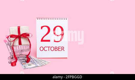 29e jour d'octobre. Une boîte cadeau dans un chariot, des dollars et un calendrier avec la date du 29 octobre sur fond rose. Automne mois, jour de t Banque D'Images