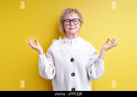 Femme âgée en lunettes de vue garder calme, namaste, isolé sur fond jaune, engagé dans la méditation de yoga, soulager la tension. Portrait de la fema ridée Banque D'Images