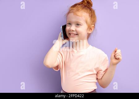 REDHEAD enfant fille communique sur téléphone mobile, sourit et parle isolé sur fond violet. Gros plan sourire enfant parlant sur smartphone. Le péché des gens Banque D'Images