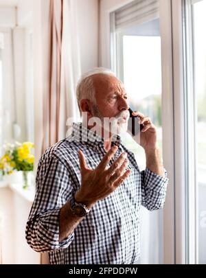Senior se tenant à côté de la fenêtre et regardant parler sur le le téléphone mobile a mis la main en place Banque D'Images