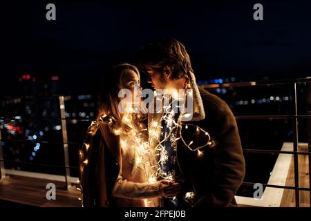 Portrait d'un beau couple avec une guirlande lumineuse sur le balcon, beau homme câlin femme magnifique, profitez de moments tendres, vacances d'hiver Banque D'Images