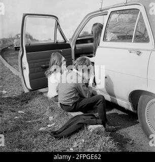 1969, historique, deux enfants assis sur le trottoir, prenant un verre, attendant devant la voiture de leur père, qui est garée sur un parking en bord de route, Wrotham Hill, Essex. Angleterre, Royaume-Uni. Banque D'Images