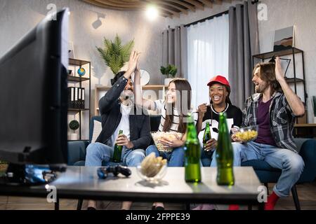 Des hommes et des femmes émotionnels qui donnent le haut cinq pour célébrer le score de l'équipe de football préférée pendant le championnat. Amis multiethniques assis sur un canapé et regardant un match à la télévision. Banque D'Images