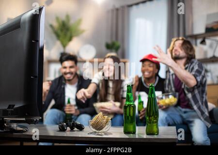 Des hommes et des femmes émotionnels qui donnent le haut cinq pour célébrer le score de l'équipe de football préférée pendant le championnat. Amis multiethniques assis sur un canapé et regardant un match à la télévision. Banque D'Images