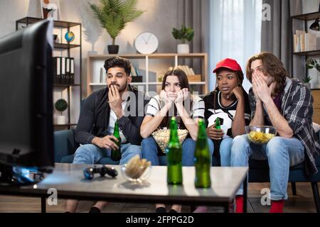 Groupe de quatre amis multiraciaux se sentent déçus tandis que leur équipe de football a perdu pendant le championnat du monde. Les jeunes hommes et les jeunes femmes regardent le match à la télévision tout en restant à la maison. Banque D'Images