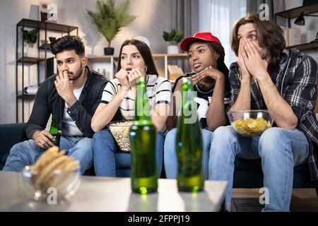 Groupe de quatre amis multiraciaux se sentent déçus tandis que leur équipe de football a perdu pendant le championnat du monde. Les jeunes hommes et les jeunes femmes regardent le match à la télévision tout en restant à la maison. Banque D'Images