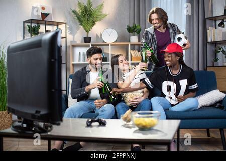 Souriant hommes et femmes faisant des acclamations avec des bouteilles de bière tout en appréciant le match de football à la télévision. Divers jeunes passent du temps ensemble pour regarder la compétition sportive à la maison. Banque D'Images