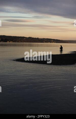 Un homme qui se framasse dans l'eau avec un poteau de pêche et de l'équipement au coucher du soleil Banque D'Images