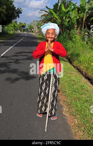 Femme balinaise âgée vêtue de sarong, hauts rouges et jaunes, serviette enveloppée autour de sa tête, debout près de la route avec les mains en position de bénédiction de prière Banque D'Images