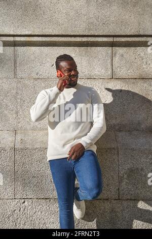 Un jeune homme africain heureux utilisant un téléphone portable et souriant appuyé contre un mur. Latino parlant au téléphone. Téléphone portable de haute qualité avec photosing et sourire contre un mur gris. Photo de haute qualité Banque D'Images