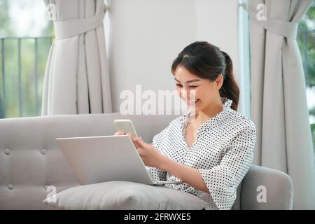 Photo d'une femme asiatique enceinte souriante utilisant un ordinateur portable et un téléphone portable tout en étant assise sur un canapé à la maison Banque D'Images