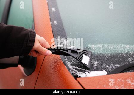 Grattoir à main. L'homme élimine la neige du pare-brise glacé de la voiture. Nettoyage de la vitre orange de l'automobile. Banque D'Images