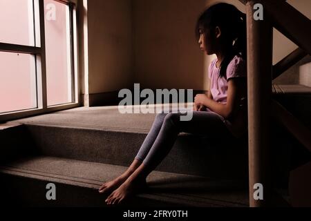 Concept de maltraitance des enfants. Une jeune fille triste et solitaire assise par un escalier à l'intérieur d'une construction avec de la lumière à travers la fenêtre en verre dans l'arrière-plan. Banque D'Images