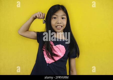 Sonnez. Une jolie jeune fille asiatique tenant une minuscule cloche en métal, l'sonnant en la secouant d'avant en arrière. Arrière-plan jaune vif. Banque D'Images