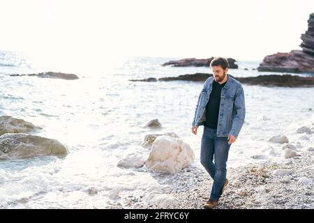 Un homme pensif en Jean et une veste en denim se promène une plage de galets près de l'eau regardant sous ses pieds sur fond de rochers Banque D'Images