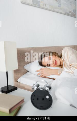 Femme souriante couchée sur le lit à proximité de livres flous et d'une alarme horloge Banque D'Images