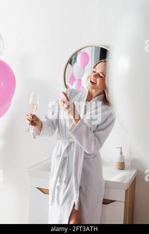 Femme souriante en peignoir prenant une photo de champagne près des ballons dans la salle de bains Banque D'Images