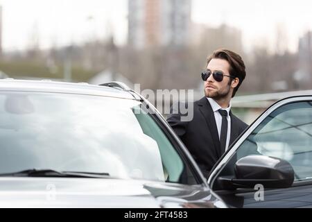 homme d'affaires barbu en costume et lunettes de soleil assis dans une voiture moderne Banque D'Images