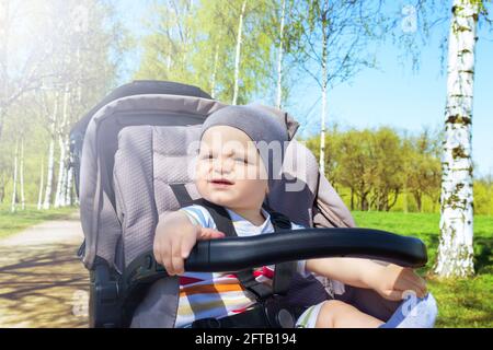 Adorable bébé caucasien heureux assis dans une poussette grise dans le parc. Jour d'été. L'enfant est attaché dans la poussette pendant la marche. Banque D'Images