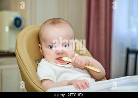 un petit enfant aux yeux bleus tient une cuillère assise dans une chaise haute. Banque D'Images