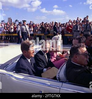 Le président John F. Kennedy et la première dame Jacqueline Kennedy se déplacent à l'arrière d'un cabriolet alors que leur cortège se rend au stade Orange Bowl à Miami, en Floride, pour une cérémonie de présentation du 2506e drapeau de la brigade d'invasion cubaine. Le président Kennedy tient sur ses genoux une grande enveloppe inscrite avec une note manuscrite, "Peech - Miami"; le maire de Miami, Robert King High, siège entre le président et Mme Kennedy (portant des gants blancs et un foulard enveloppé sous son menton). Sur le siège avant de la voiture (de gauche à droite) : agent du service secret de la Maison Blanche, Floyd Boring.