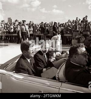 Le président John F. Kennedy et la première dame Jacqueline Kennedy se déplacent à l'arrière d'un cabriolet alors que leur cortège se rend au stade Orange Bowl à Miami, en Floride, pour une cérémonie de présentation du 2506e drapeau de la brigade d'invasion cubaine. Le président Kennedy tient sur ses genoux une grande enveloppe inscrite avec une note manuscrite, "Peech - Miami"; le maire de Miami, Robert King High, siège entre le président et Mme Kennedy (portant des gants blancs et un foulard enveloppé sous son menton). Sur le siège avant de la voiture (de gauche à droite) : agent du service secret de la Maison Blanche, Floyd Boring. Banque D'Images