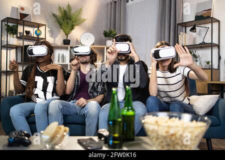 Groupe de quatre amis multi-ethniques utilisant un casque sans fil pour découvrir la réalité virtuelle. Des hommes et des femmes joyeux assis sur un canapé confortable et jouant à des jeux. Concept de technologie. Banque D'Images