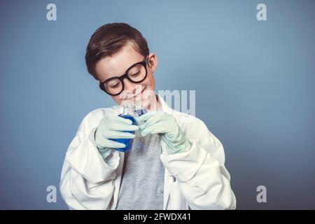 cool garçon d'école avec des lunettes noires épaisses est habillé comme scientifique à couche blanche et expérimentant avec des liquides bleus dans avant de l'arrière-plan bleu Banque D'Images