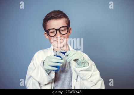 cool garçon d'école avec des lunettes noires épaisses est habillé comme scientifique à couche blanche et expérimentant avec des liquides bleus dans avant de l'arrière-plan bleu Banque D'Images