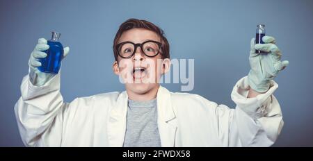 cool garçon d'école avec des lunettes noires épaisses est habillé comme scientifique à couche blanche et expérimentant avec des liquides bleus dans avant de l'arrière-plan bleu Banque D'Images