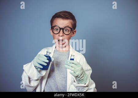 cool garçon d'école avec des lunettes noires épaisses est habillé comme scientifique à couche blanche et expérimentant avec des liquides bleus dans avant de l'arrière-plan bleu Banque D'Images