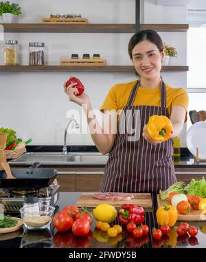 La jeune femme de ménage se tient souriante, tient le poivron rouge et jaune avec les deux mains. En proposant le jaune à l'avant. Le comptoir de cuisine plein de Banque D'Images