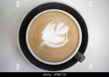 Café, cappuccino avec mousse de lait en forme de feuille dans une tasse blanche sur une soucoupe noire sur un fond de table blanc. Vue de dessus. Banque D'Images