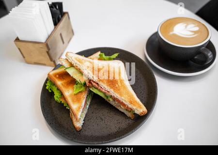 Petit déjeuner d'affaires le matin : sandwich au saumon et cappuccino dans un élégant plat noir sur une table blanche Banque D'Images