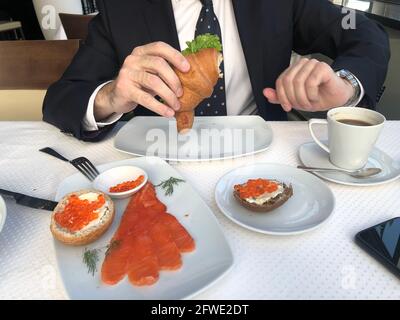 Homme dans un costume d'affaires, cravate et chemise blanche est de prendre le petit déjeuner, pause-café, une tasse de café, croissant avec saumon et salade verte, sandwichs avec Banque D'Images