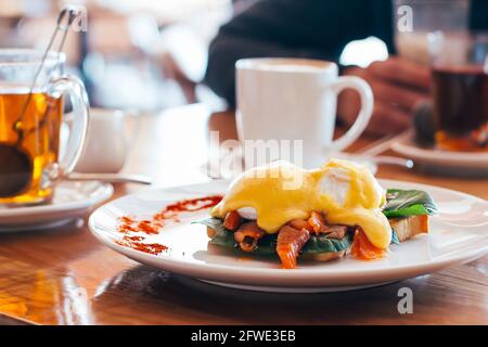 Œuf poché sur un morceau de pain avec du saumon et des épinards sur l'assiette, mugs avec café et thé sur une table en bois dans le café. Concept petit déjeuner matin Banque D'Images