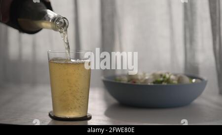 man verse de la bière au gingembre provenant d'une bouteille de verre dans un verre à gobelet avec salade dans un bol sur une table en chêne blanc avec coucher de soleil photo grand angle clair Banque D'Images