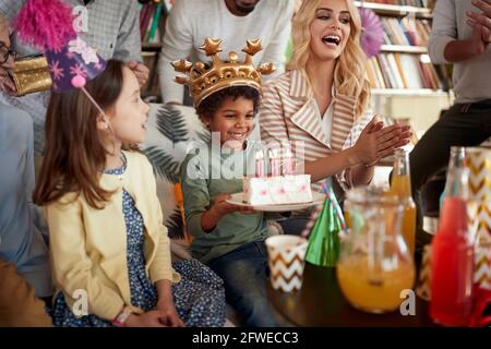 Un petit garçon heureux tenant un gâteau à sa fête d'anniversaire dans une atmosphère festive à la maison avec la famille et les amis. Famille, célébration, ensemble Banque D'Images