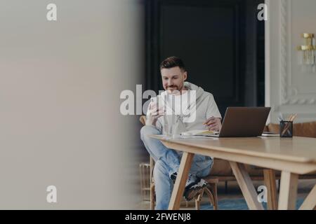 Joyeux jeune homme entrepreneur assis près d'une table en bois et en utilisant smartphone tout en travaillant à distance sur un ordinateur portable depuis chez vous Banque D'Images